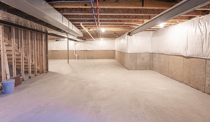 A spacious, well-lit basement with freshly waterproofed concrete floors, white moisture-barrier wall panels, and exposed wooden ceiling joists